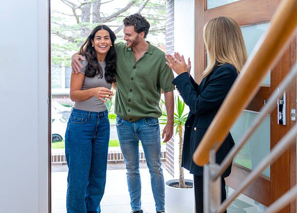 Real estate agent or landlord giving house keys to a young couple at the front door of their new house. Everyone is happy and smiling.